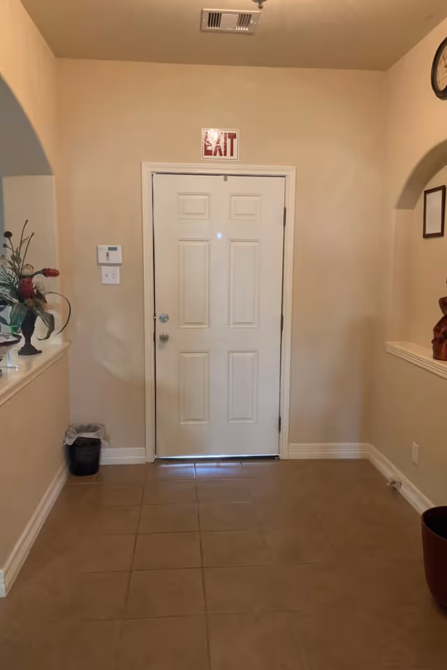 Interior view of a hallway with a closed white door marked with an EXIT sign above it. The hallway has beige walls and tiled floor. On the left side, there is a small trash bin and a decorative vase with flowers on a ledge. On the right side, there is a clock on the wall and a framed document.