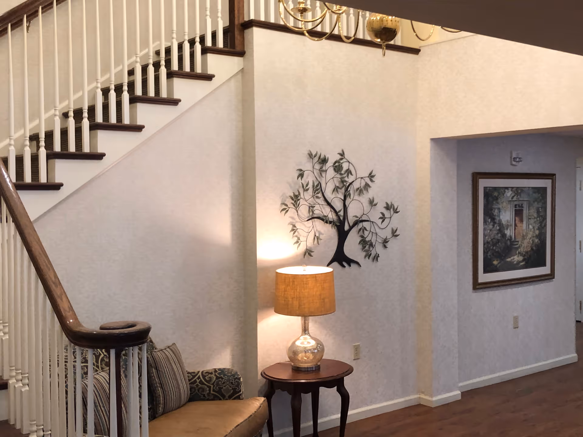 Interior view of a senior living facility showing a staircase with white railings and dark wooden steps. Below the staircase is a cushioned chair with patterned upholstery and striped pillows. Next to the chair is a small wooden side table with a lamp that has a beige shade. On the wall above the lamp is a decorative metal tree sculpture. Further down the hallway, there is a framed painting on the wall. The floor is wooden, and the walls are light-colored.
