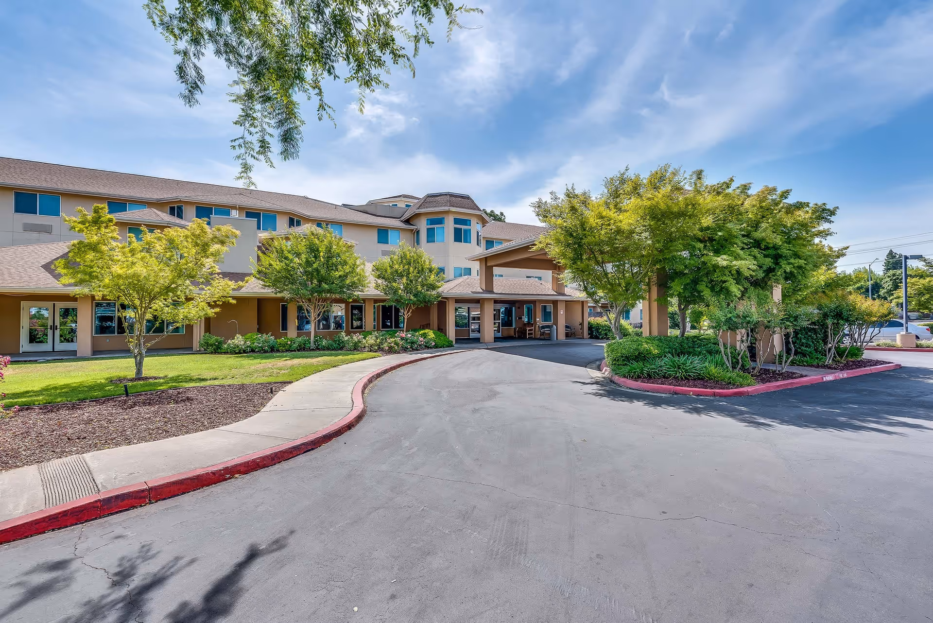 Exterior view of Solstice Senior Living at Lodi showing a multi-story building with beige walls and a covered entrance. The driveway curves in front of the entrance, surrounded by well-maintained landscaping including green trees, bushes, and grass under a partly cloudy blue sky.