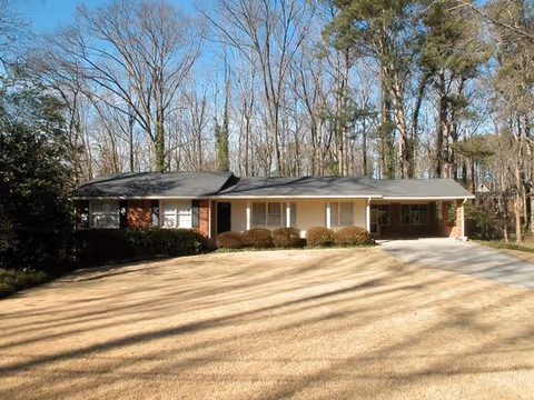 Single-story brick house with a large front yard and driveway, surrounded by tall trees with no leaves under a clear blue sky.