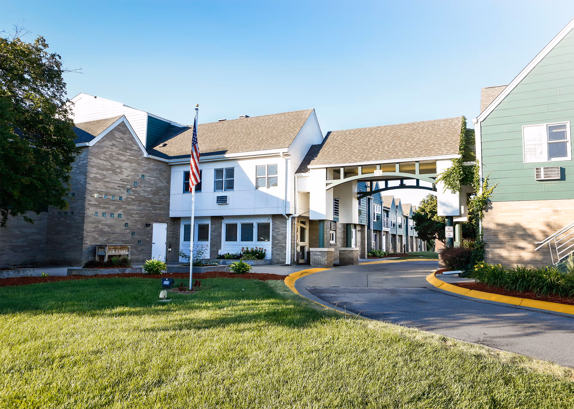 Exterior view of Hickory Villa Assisted Living facility showing a driveway passing under a covered archway connecting two buildings, with well-maintained green lawns, an American flag on a flagpole, and clear blue sky.