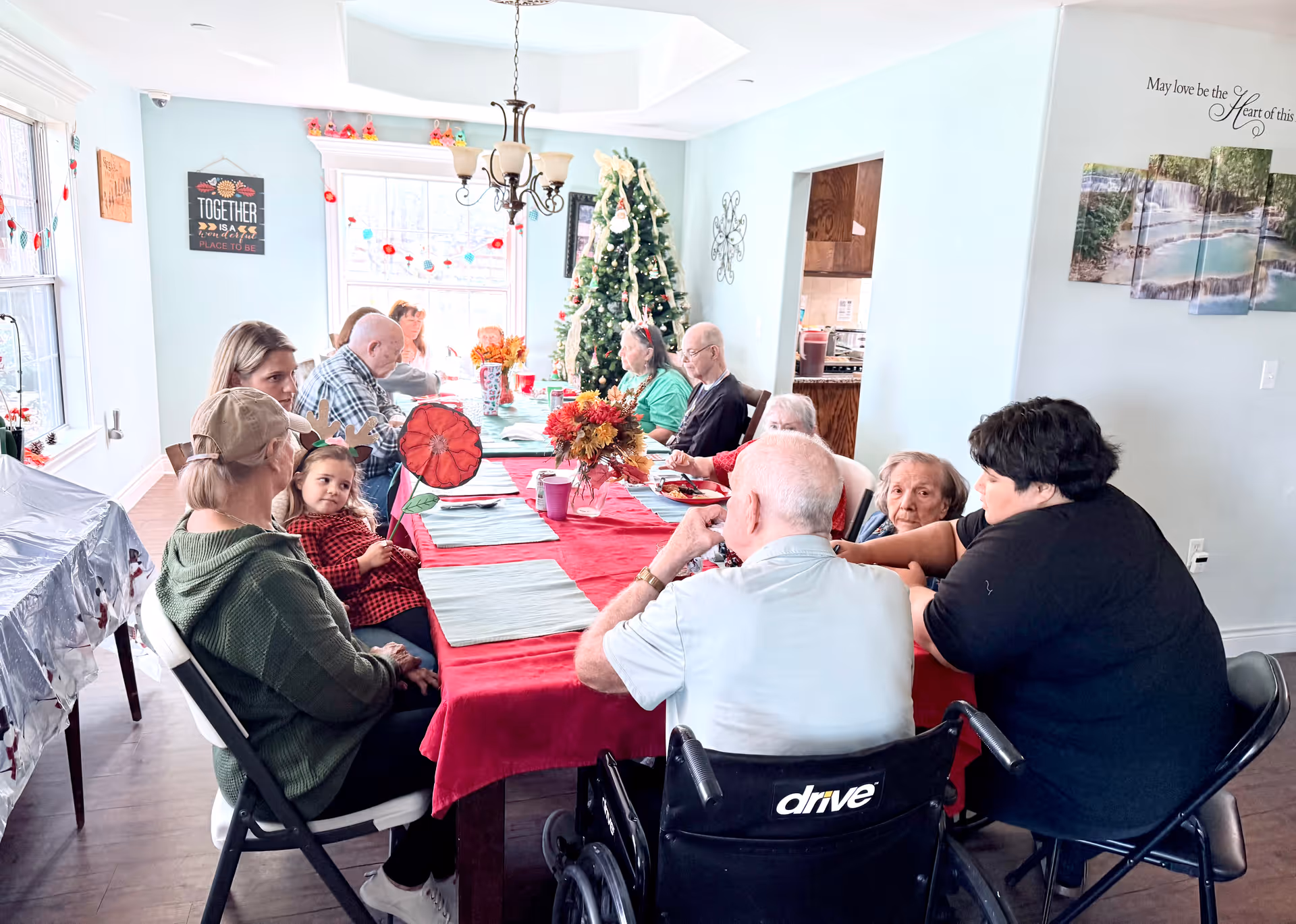 A group of elderly people and a few younger adults sitting around a long dining table covered with a red tablecloth and light blue placemats. The room is decorated with a Christmas tree and festive decorations. The setting appears to be a communal dining area with large windows letting in natural light.
