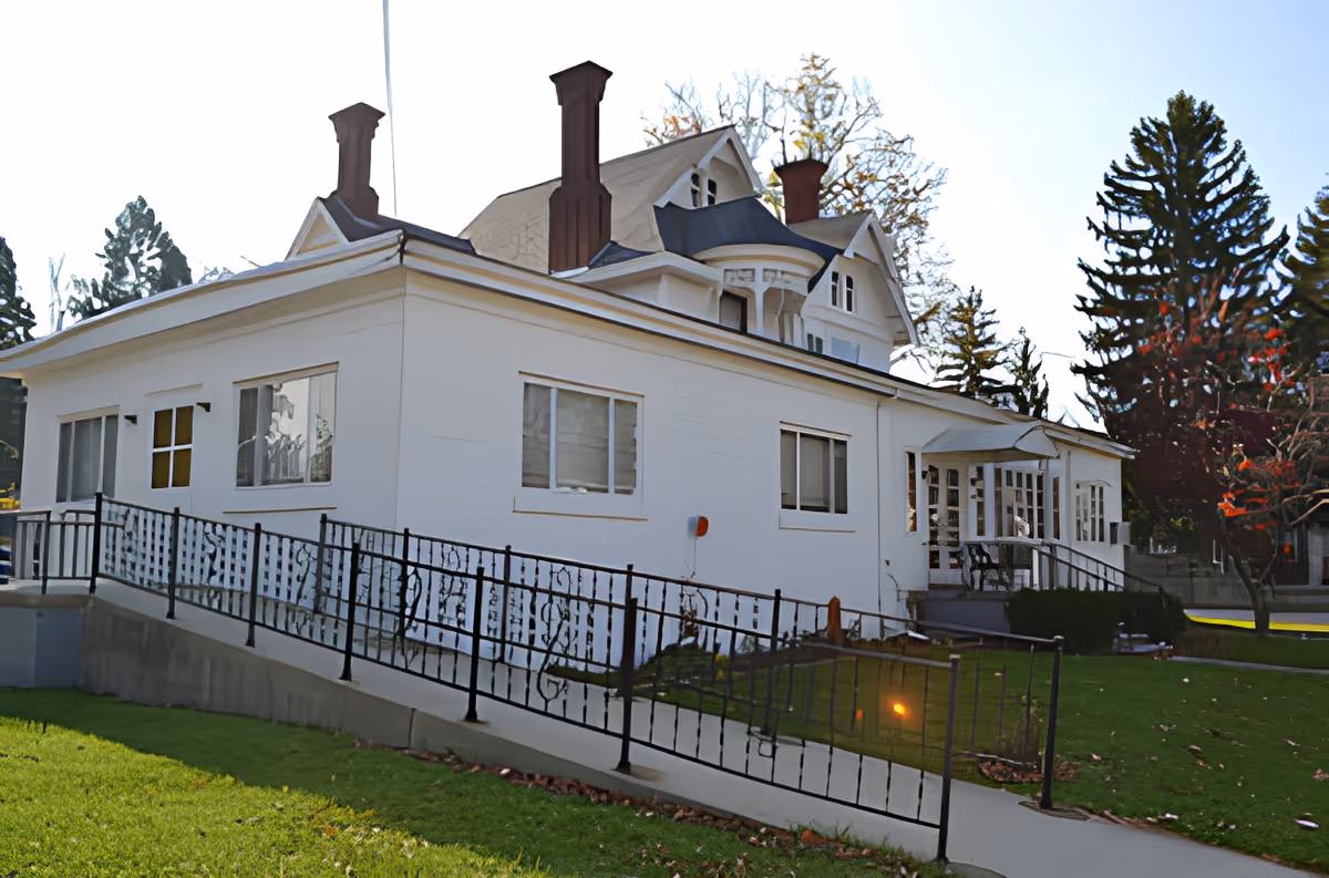 Exterior view of a white multi-story building with several chimneys and a ramp with black metal railings leading to the entrance. The building is surrounded by green grass and trees with some autumn foliage.