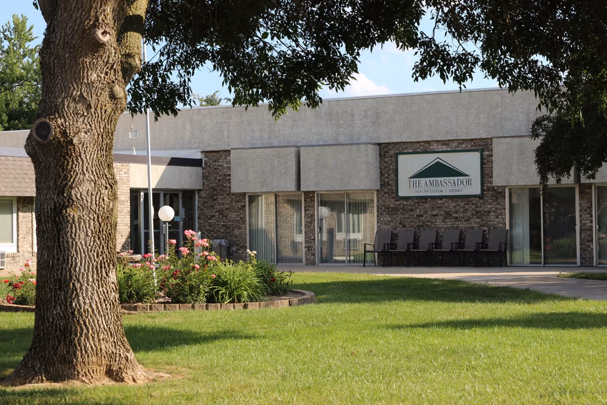 Exterior view of a single-story building with a sign that reads 'The Ambassador Health System Sidney'. The building has large windows and a row of chairs outside. In the foreground, there is a large tree, green grass, and a flower bed with pink flowers.