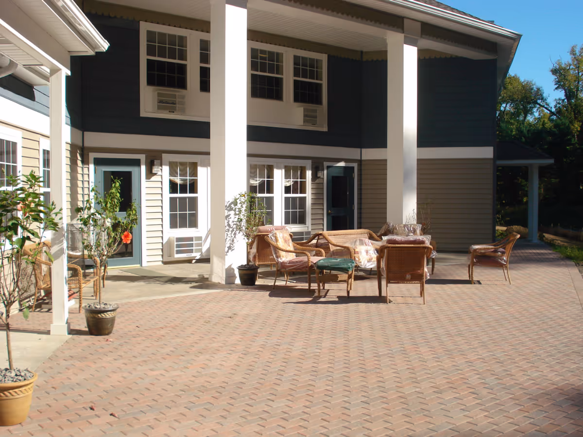 Sunlit outdoor patio courtyard with wicker chairs, potted plants, and columns in front of a two-story building.