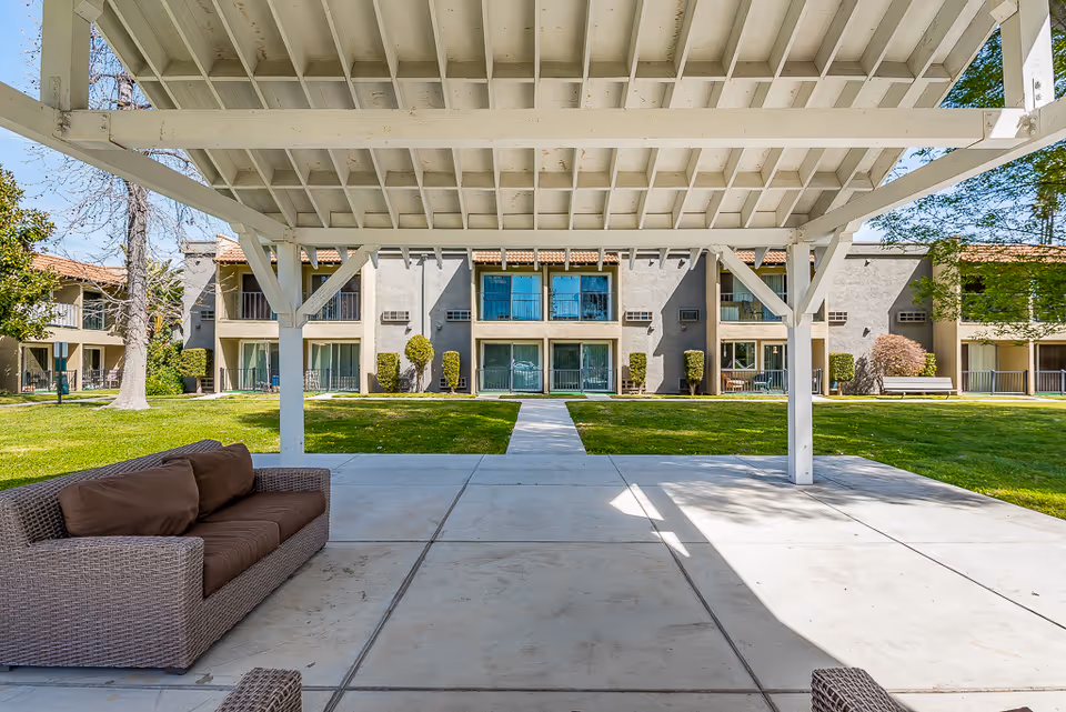View from under a white wooden pergola with a brown cushioned outdoor sofa on a concrete patio, overlooking a green lawn and a two-story senior living facility building with balconies and large windows.