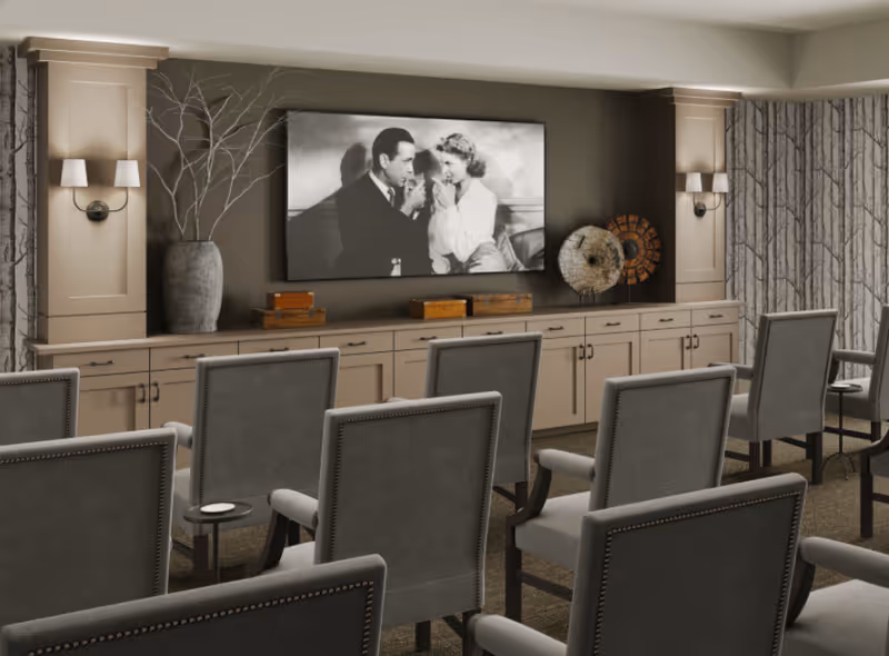 A cozy common area with rows of gray upholstered chairs facing a large flat-screen TV mounted on a taupe wall. The TV displays a black and white image of a man and woman. Below the TV is a long cabinet with decorative items including a large vase with branches and two circular art pieces. The room has soft lighting from wall sconces and patterned curtains on the windows.