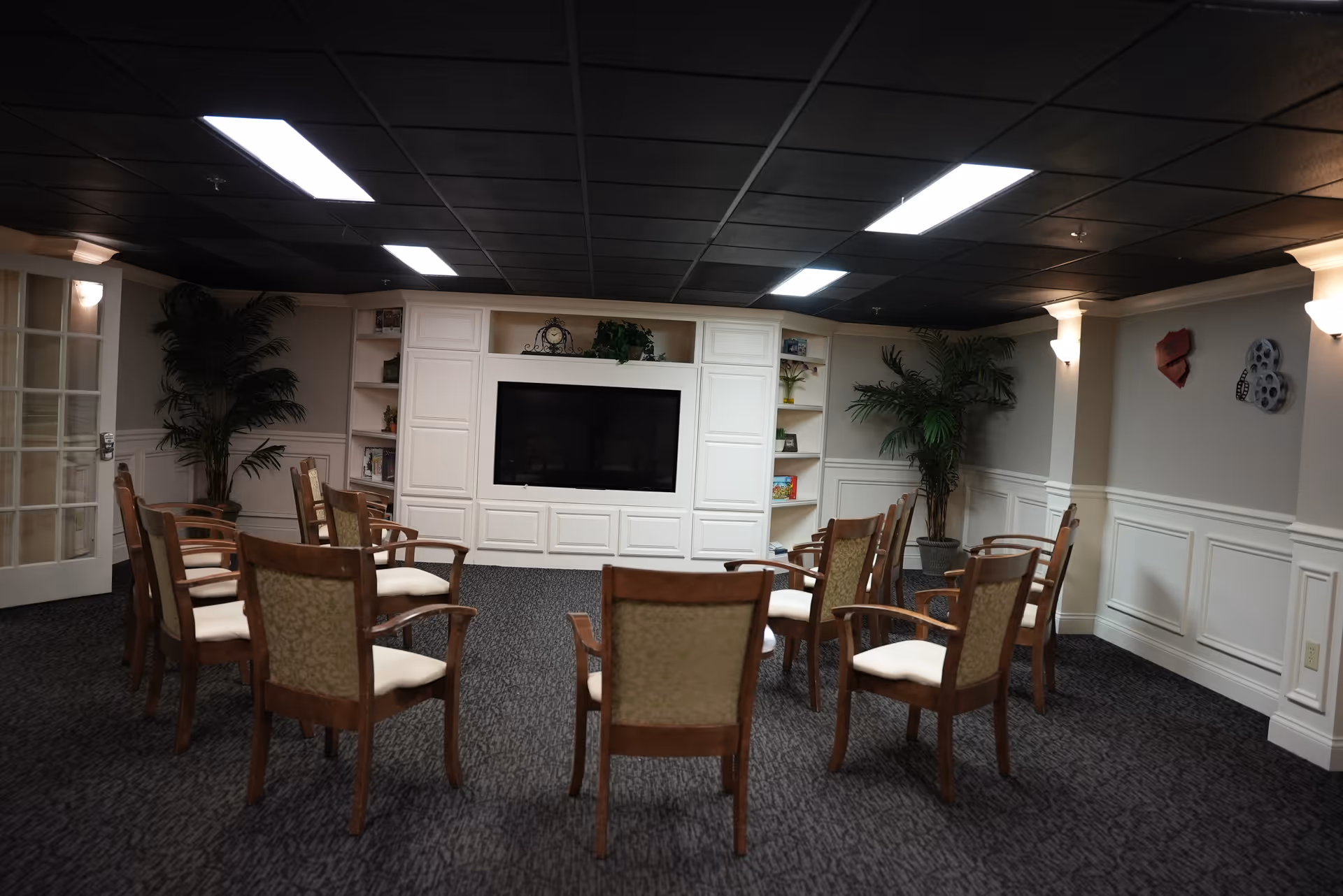 A meeting or activity room with wooden chairs arranged in a circle on a carpeted floor. The room has a large flat-screen TV mounted on a white built-in cabinet with shelves holding books and decorative items. There are two large potted plants in the corners, wall sconces providing lighting, and decorative wall art on the right wall.
