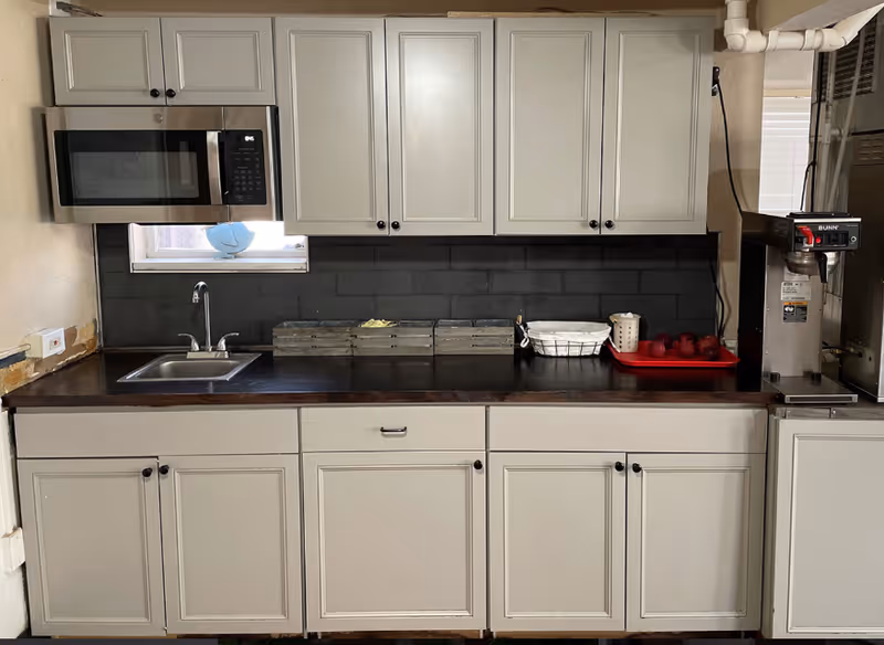 A kitchen countertop with light cabinets, a sink, microwave, dark backsplash, and a coffee machine with a red tray of fruit.