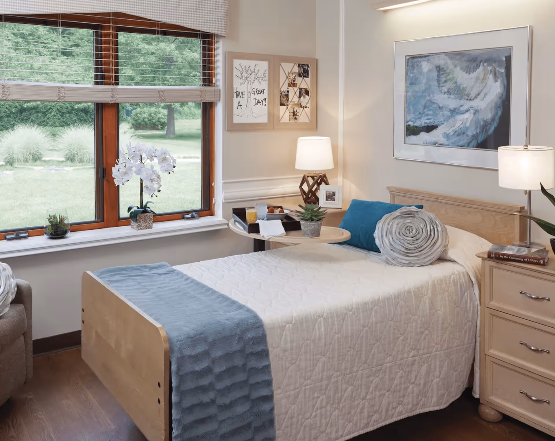 A cozy bedroom with a single bed covered in a white quilt and a blue throw blanket. The bed has a blue pillow and a rolled gray blanket. Next to the bed is a wooden nightstand with a lamp and a book. A small round table holds a tray with breakfast items and a small plant. The room has a large window with a view of green trees and grass outside. The walls are decorated with framed artwork and a lamp on a wooden base.