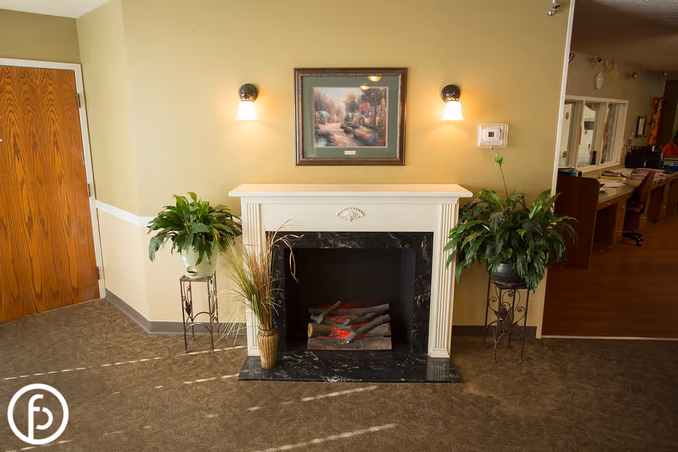 Decorative fireplace with potted plants and a framed painting above, with a reception area visible to the right.