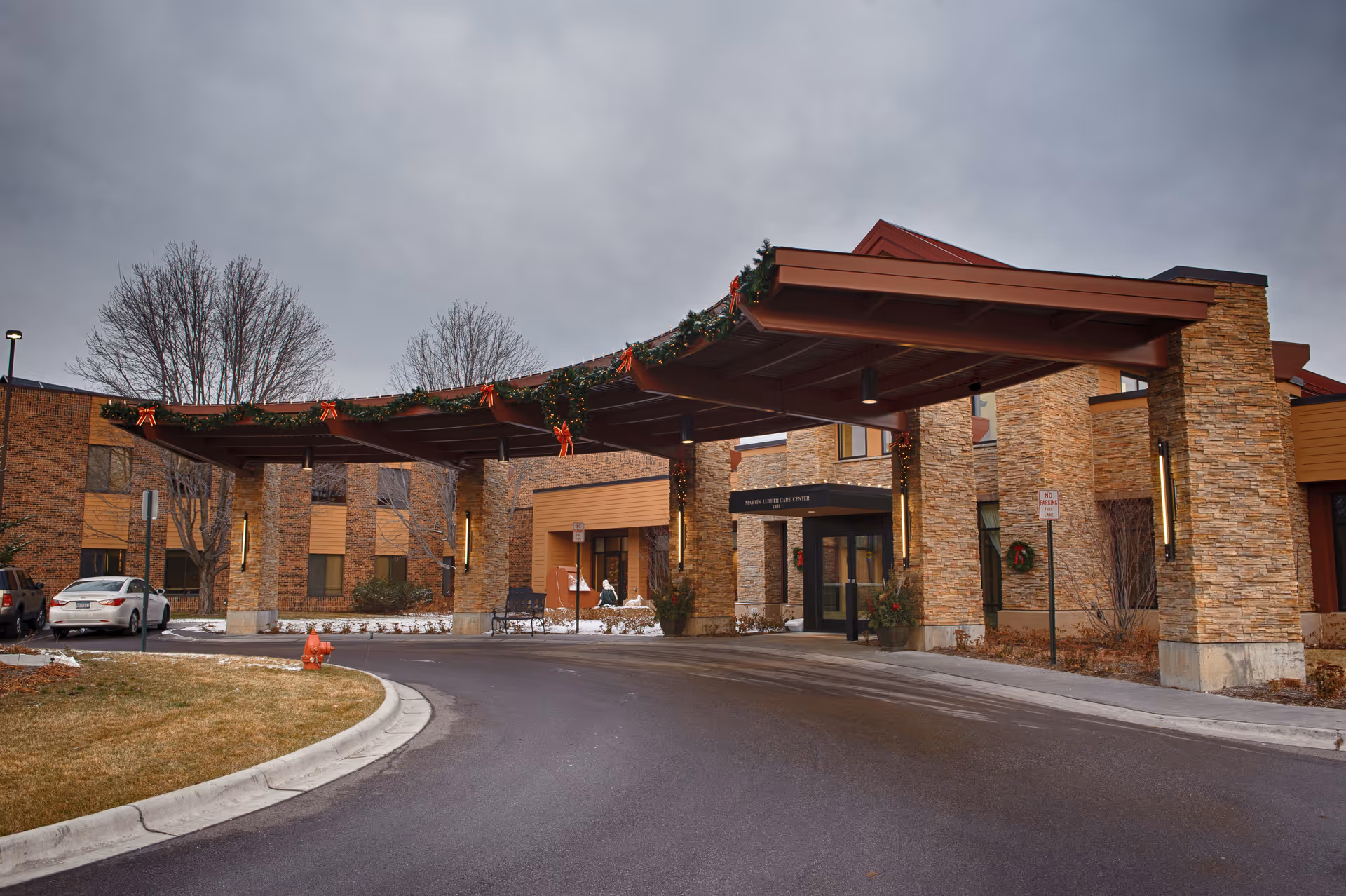 Front entrance of a senior living building with a covered porte-cochere supported by stone pillars decorated for the holidays.