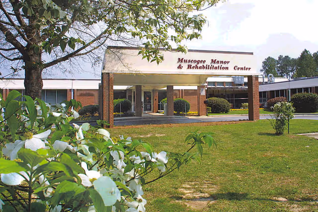 Exterior view of Muscogee Manor & Rehabilitation Center showing the entrance with a covered drop-off area supported by brick columns. The building is surrounded by green grass, bushes, and trees with white flowers in the foreground under a partly cloudy sky.