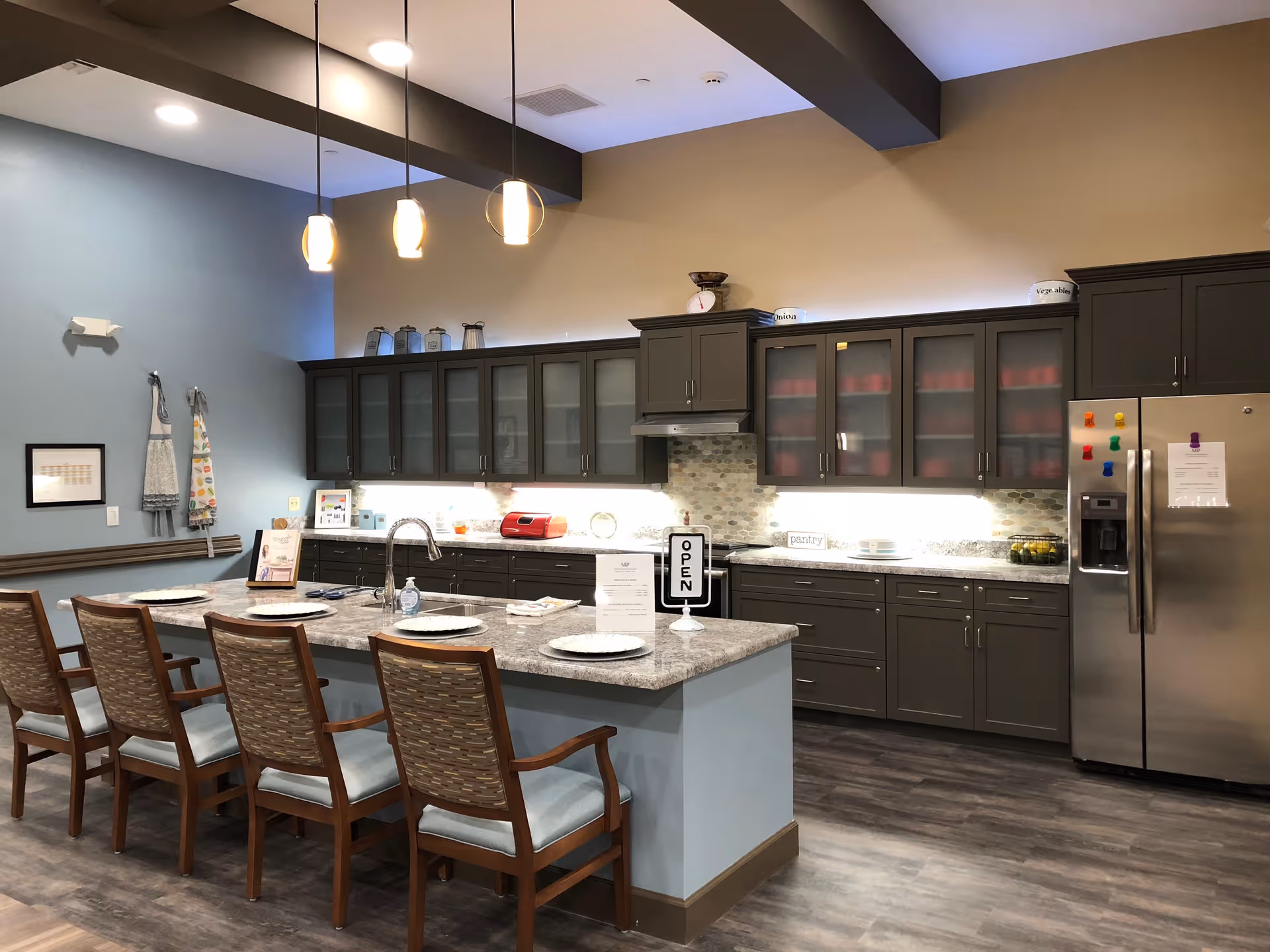 Modern communal kitchen with a large island, pendant lights, bar seating, dark cabinets, and a stainless steel refrigerator.