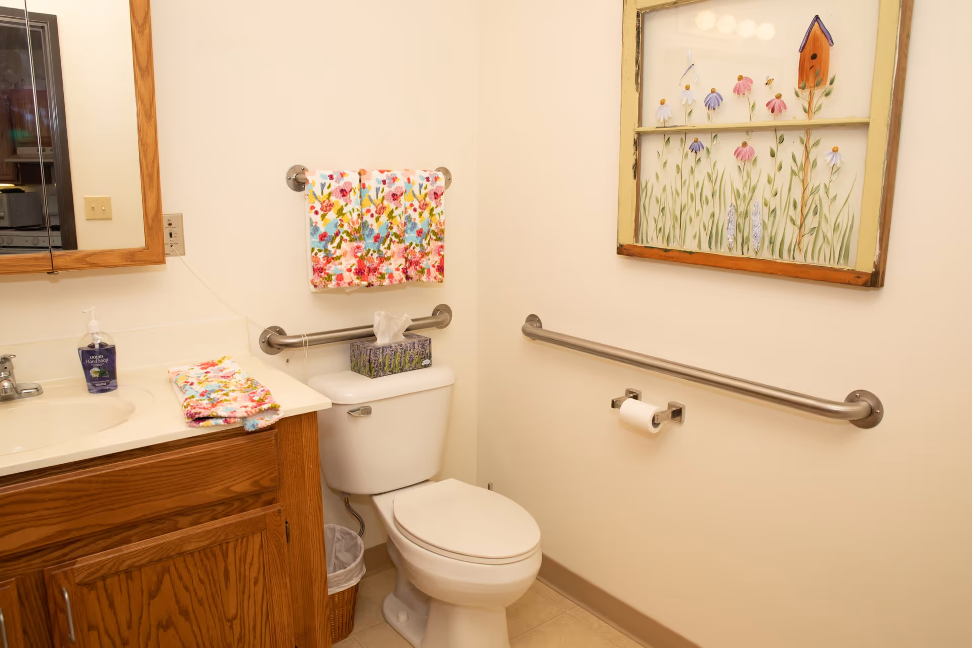 A clean bathroom with a white toilet, wooden vanity with a sink, a mirror above the sink, colorful floral towels hanging on a towel rack, a tissue box on the toilet tank, and a decorative framed artwork of flowers and a birdhouse on the wall. There are metal grab bars installed on the walls for support.