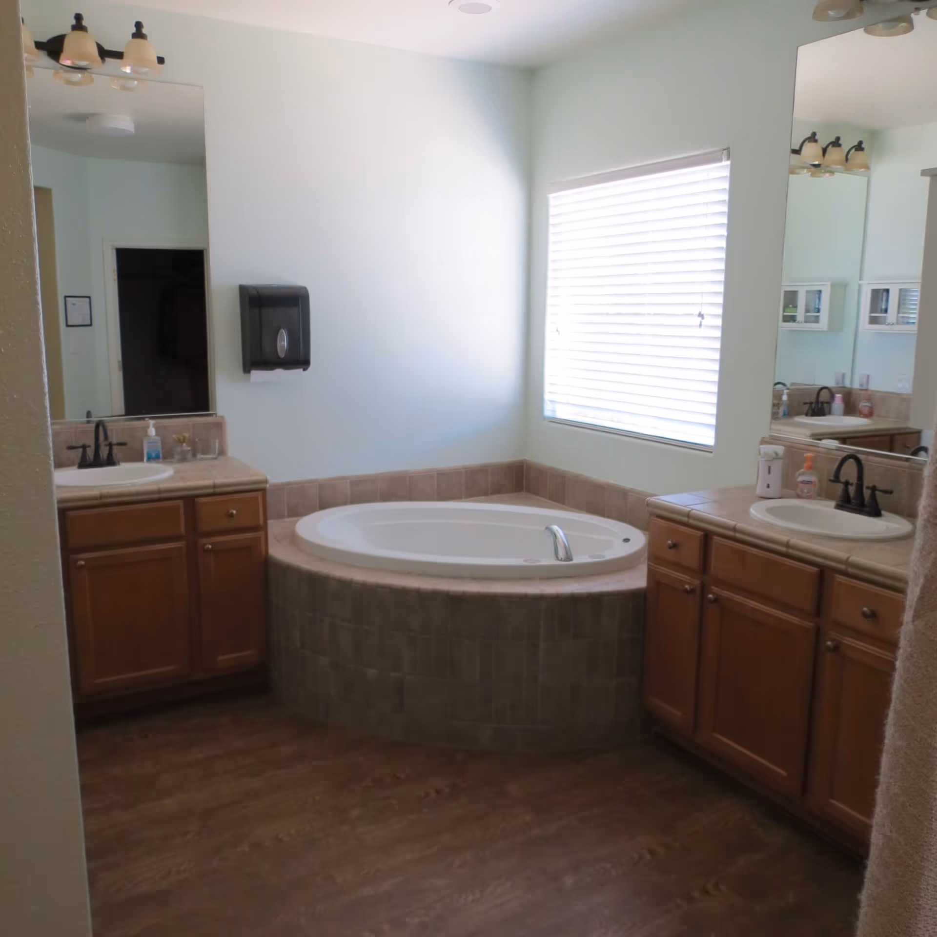 A bathroom with a large corner bathtub surrounded by beige tiles. There are two wooden vanities with sinks on either side of the bathtub, each with a mirror above. A window with white blinds is on the wall above the bathtub, and a paper towel dispenser is mounted on the wall to the left. The floor has a wood-like finish.