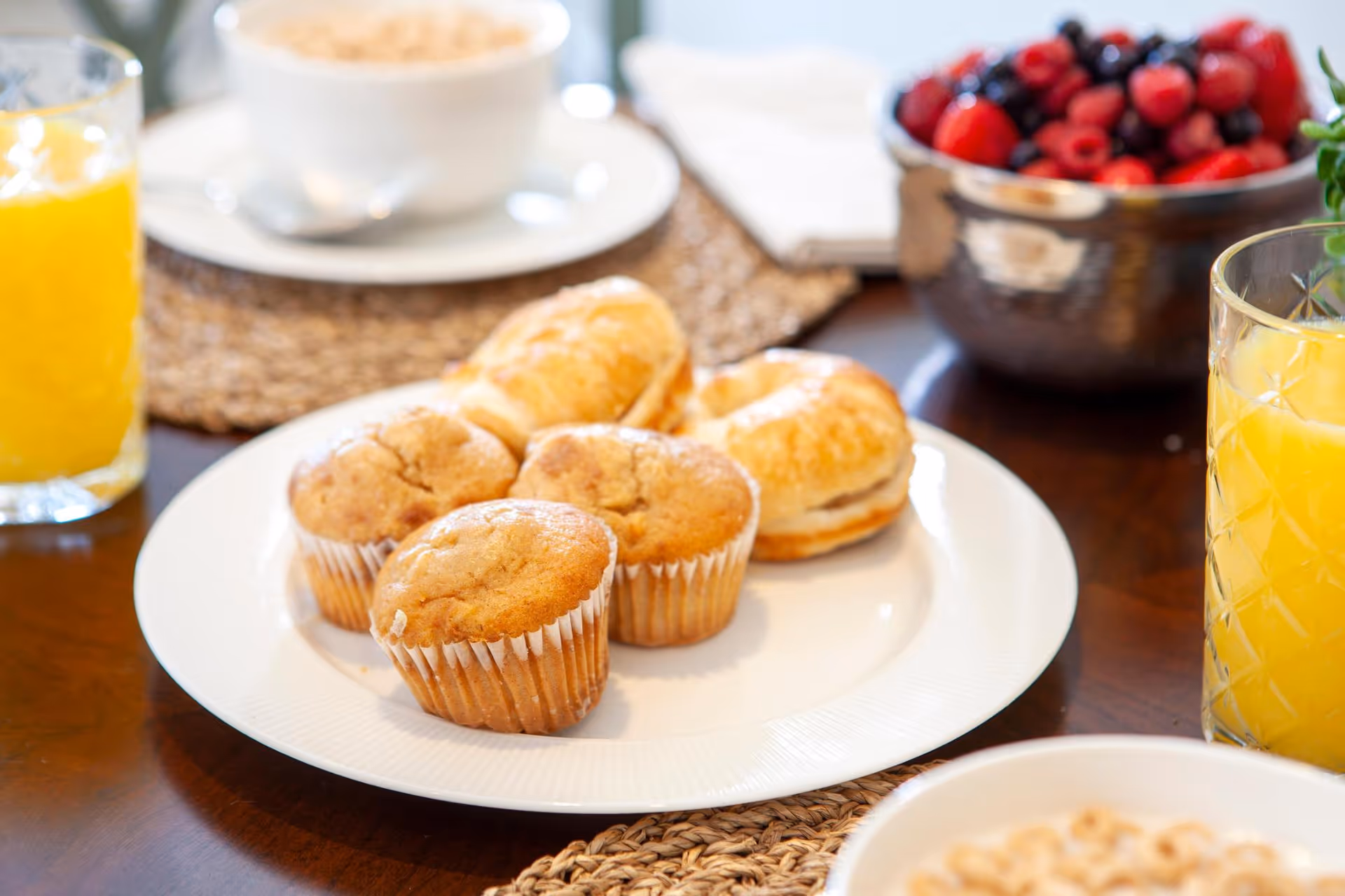 Close-up of a breakfast table with muffins and bagels on a white plate, glasses of orange juice, a bowl of berries and a cereal bowl.
