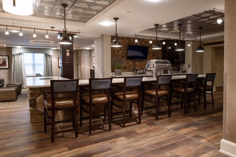 Interior view of a senior living facility common area featuring a long kitchen island with eight high chairs, pendant lighting above, a mounted flat-screen TV on a stone accent wall, and a seating area with windows and curtains in the background.