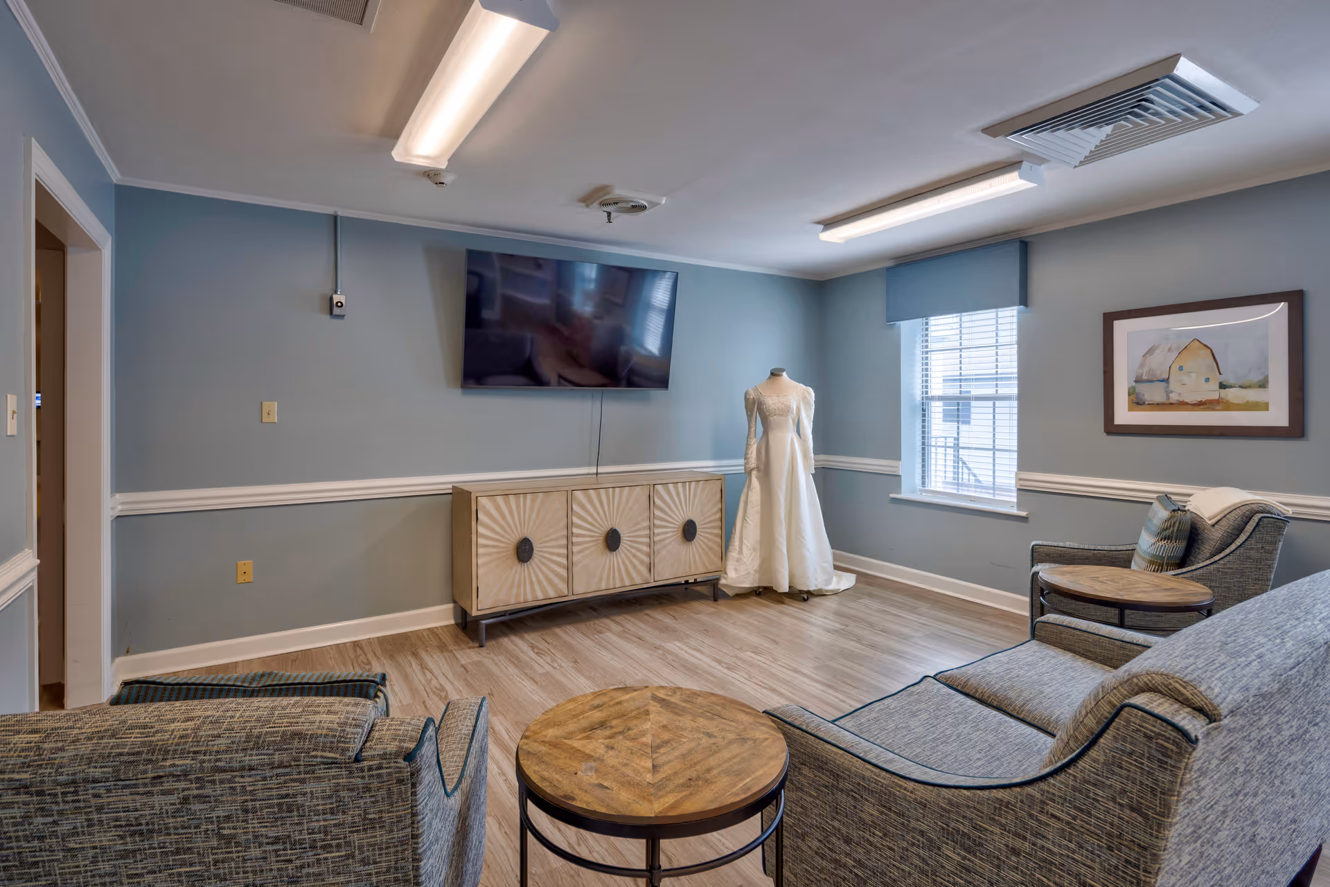 A living room with light blue walls and wood flooring featuring a flat-screen TV mounted above a decorative cabinet. There are two upholstered armchairs and a matching sofa arranged around a round wooden coffee table. A mannequin dressed in a white wedding gown stands near a window with blinds and a blue valance. A framed painting of a barn hangs on the wall beside the window.