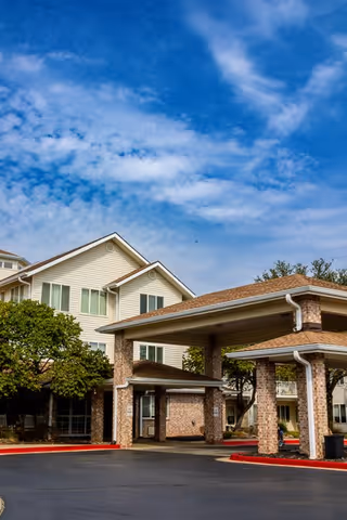 Exterior view of a senior living facility with a covered entrance supported by brick pillars, surrounded by trees and a clear blue sky with some clouds.
