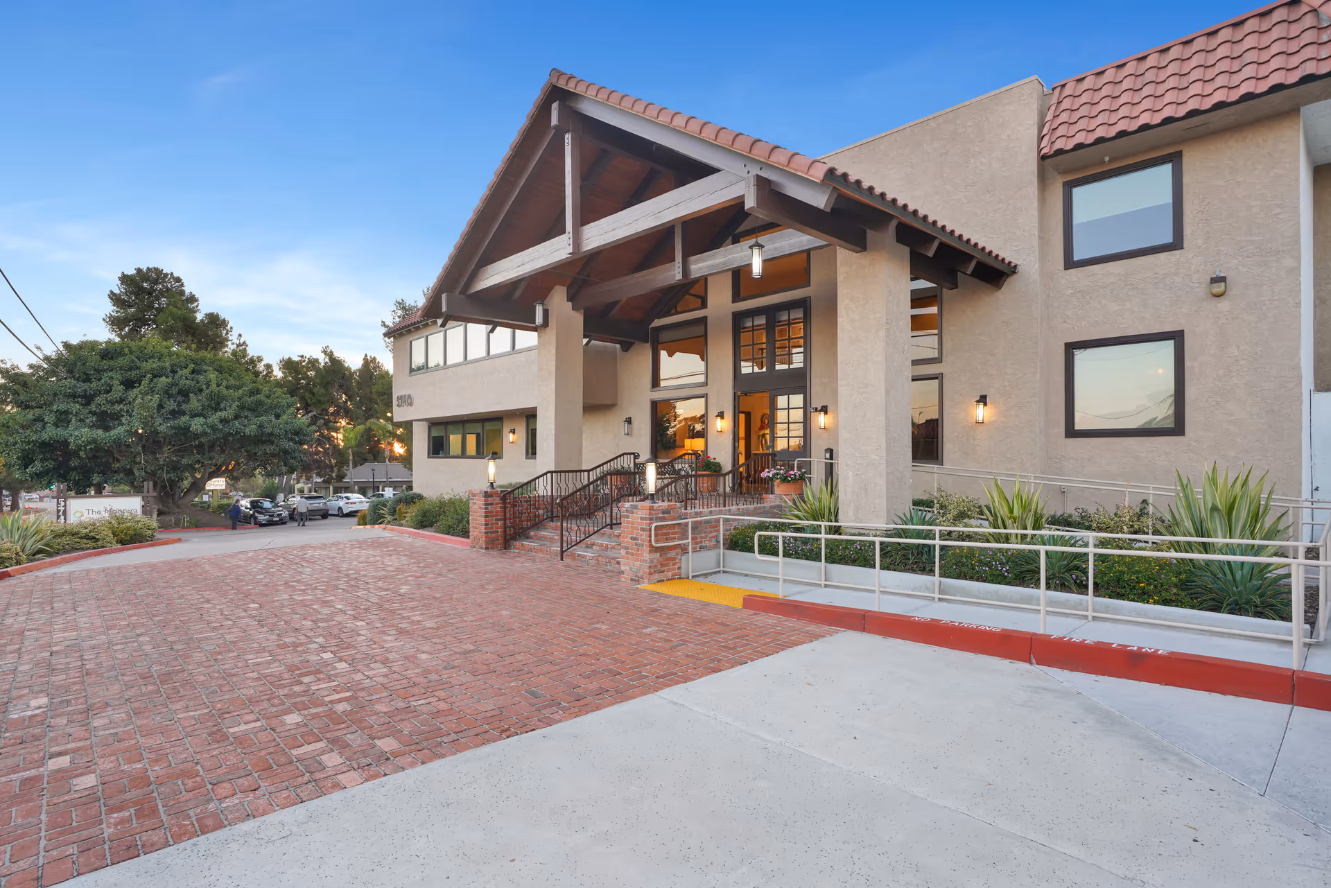Exterior view of The Montera senior living facility showing the entrance with a covered porch, brick steps, a wheelchair ramp, and surrounding greenery under a clear blue sky.
