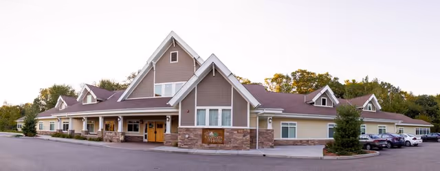 Exterior view of a single-story assisted living facility building with a peaked roof, stone and siding facade, multiple windows, and a parking lot with several cars. The building is surrounded by trees and greenery.