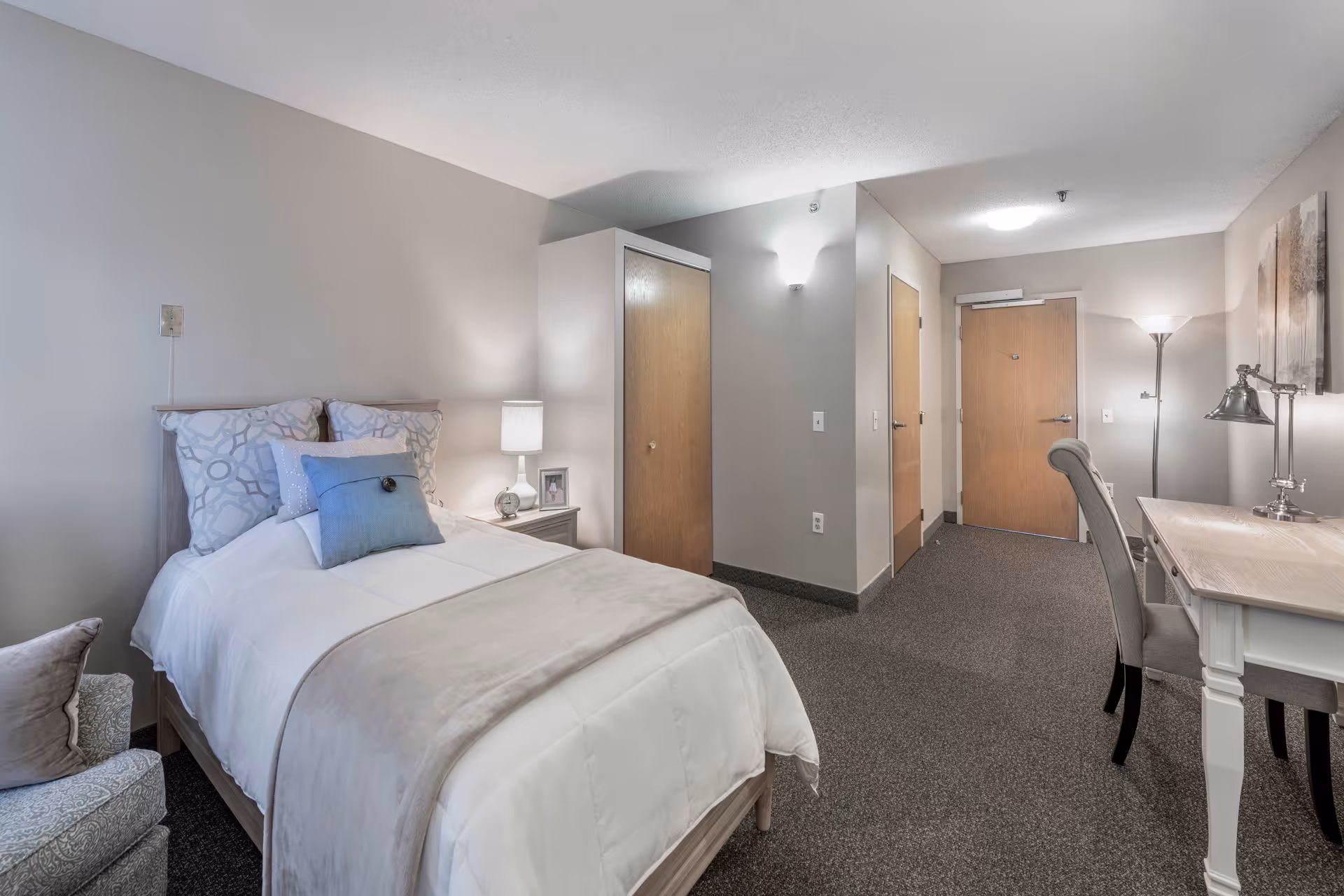 Bright, neatly furnished senior living bedroom with a single bed, bedside table and lamp, a desk and chair, and an entry door.