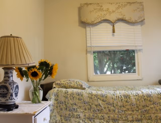 Sunlit single bedroom with a floral bedspread, a nightstand holding a lamp and vase of sunflowers, and a window with blinds.