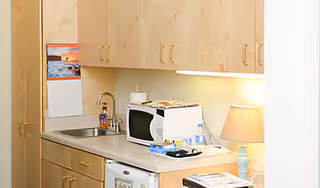 A small kitchenette area with light wood cabinets, a stainless steel sink, a white microwave, a table lamp with a blue base, and various items including a calendar, paper towels, and some papers on the countertop.