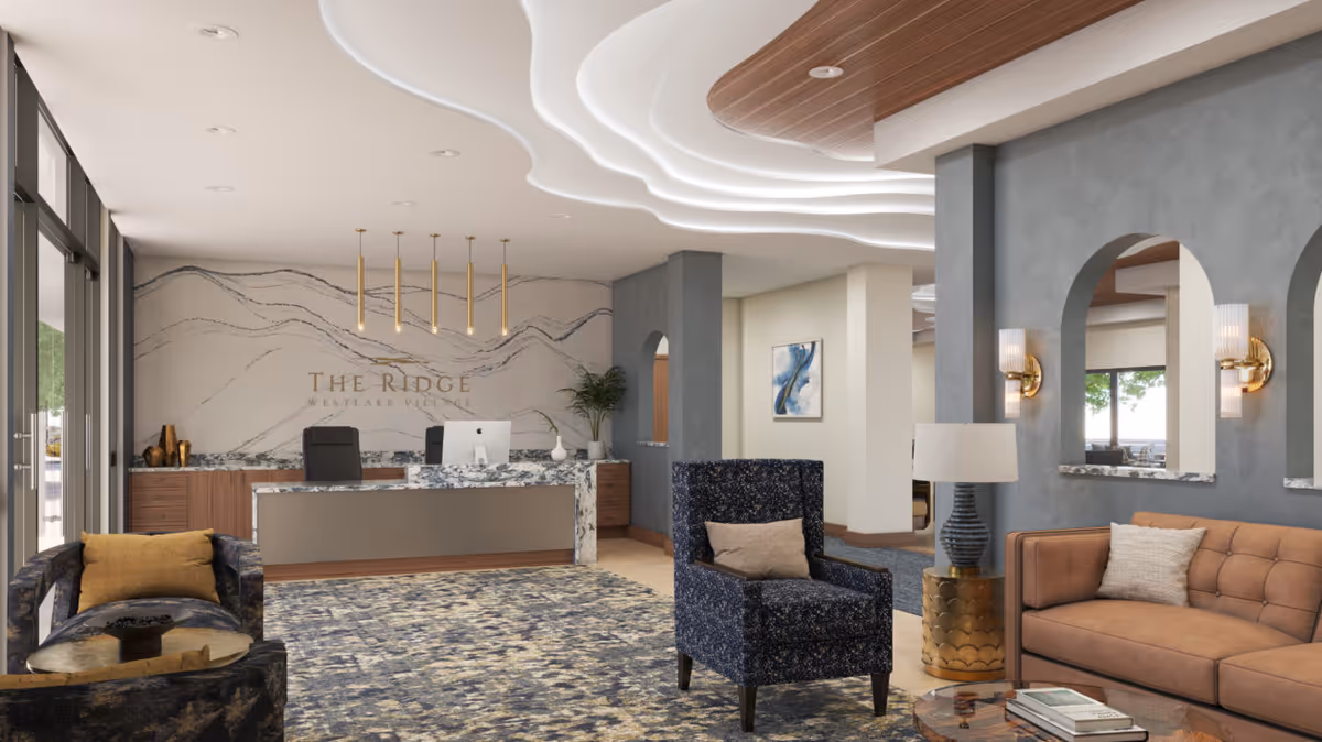 Bright reception lobby with a marble front desk, seating area of chairs and a sofa, decorative ceiling lighting, and a wall sign reading 'The Ridge'.