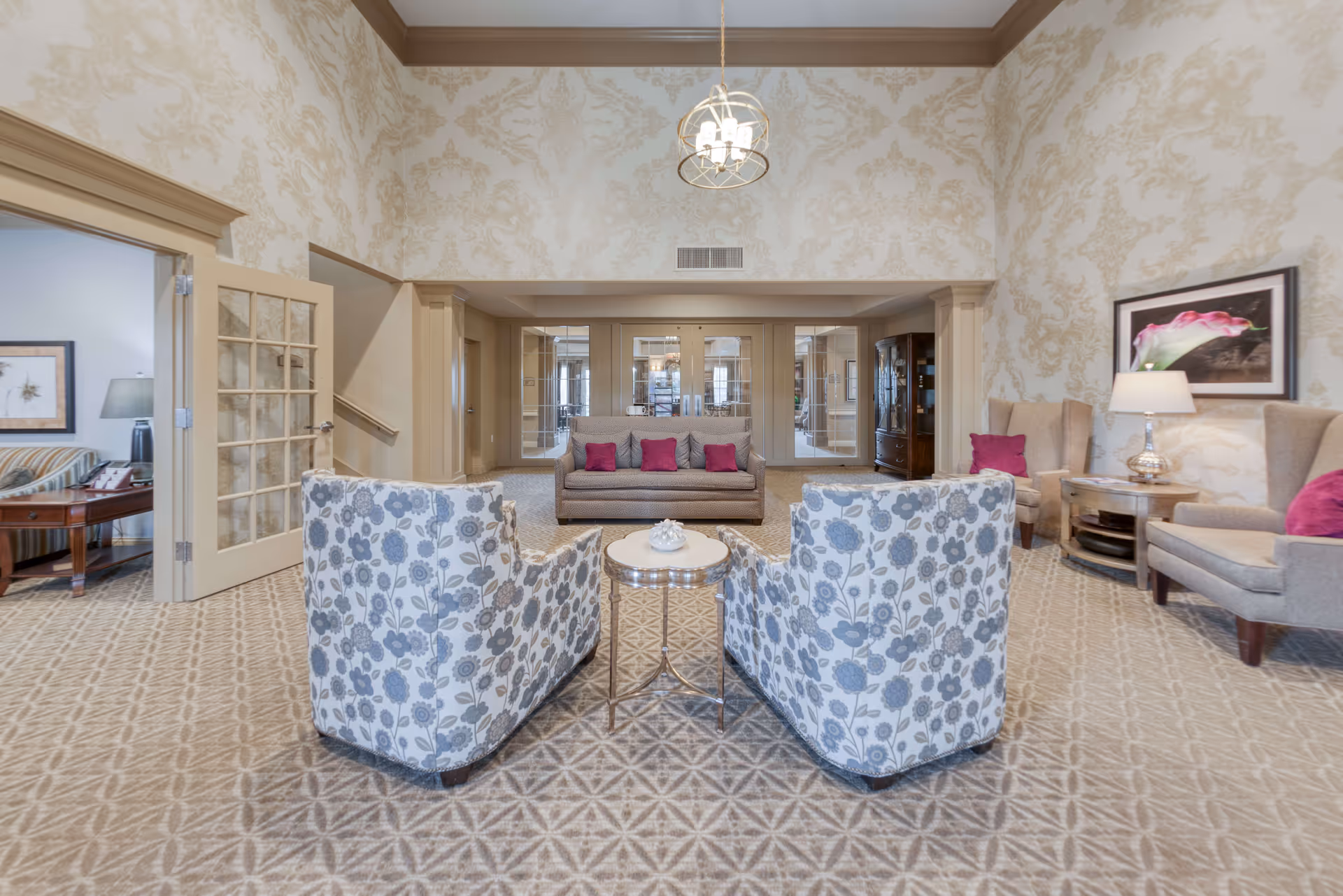 A spacious and elegantly decorated living room with high ceilings and patterned wallpaper. The room features two floral-patterned armchairs facing a small round table, a gray sofa with pink cushions, and two beige armchairs with pink cushions. There is a framed flower picture on the wall and a table lamp on a side table. The room has beige carpeting with a geometric pattern and a chandelier hanging from the ceiling. A doorway with glass-paneled double doors leads to another room with a striped sofa and a lamp.