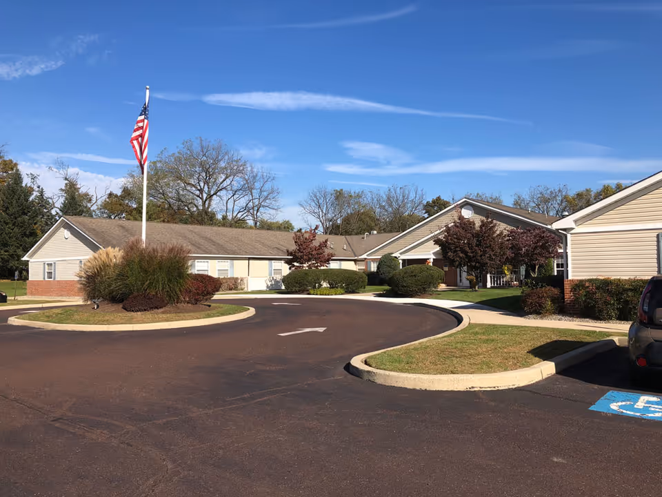 Exterior view of Arden Courts - ProMedica Memory Care Community in Warminster, showing a single-story building with beige siding and brick accents, surrounded by landscaped bushes and trees. An American flag is flying on a flagpole in the center of a circular driveway under a clear blue sky.