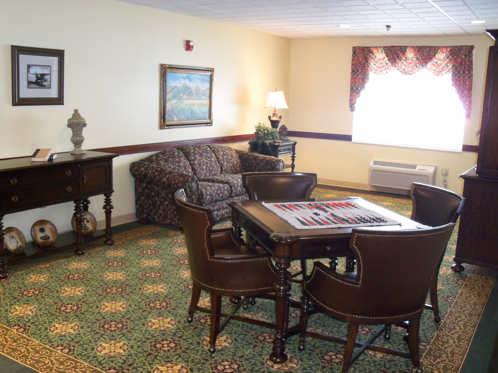 A cozy sitting area in a senior living facility with a patterned green carpet, a floral upholstered couch, a wooden table with a checkerboard game on top, and four leather chairs around the table. The room has light yellow walls with framed artwork, a side table with a lamp and plants, and a window with floral curtains letting in bright light.