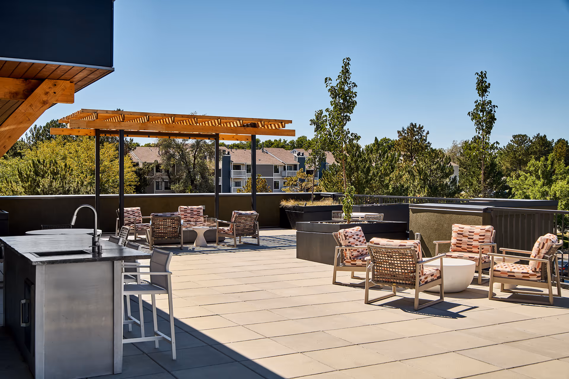 Rooftop outdoor patio with seating, a pergola, and an outdoor kitchen overlooking trees and nearby buildings.
