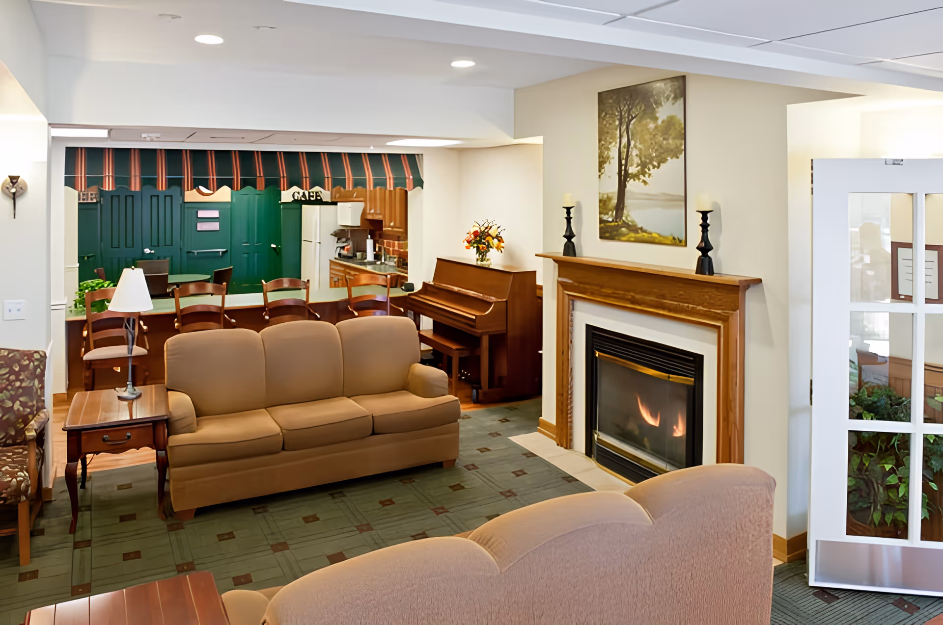 A cozy common living room with sofas facing a fireplace, a piano, and a dining area visible in the background.