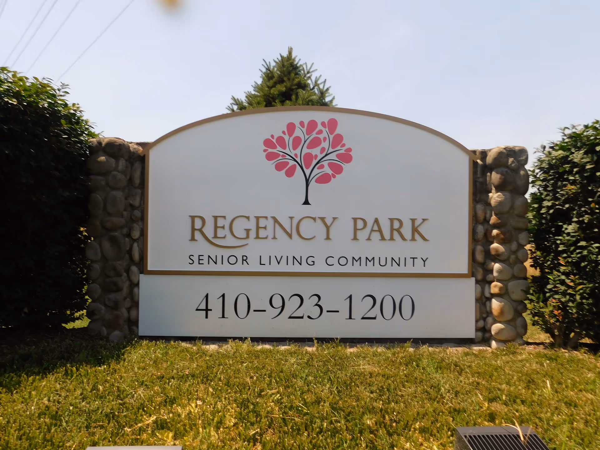 Outdoor sign for Regency Park Senior Living Community with a decorative tree logo and phone number between stone pillars.