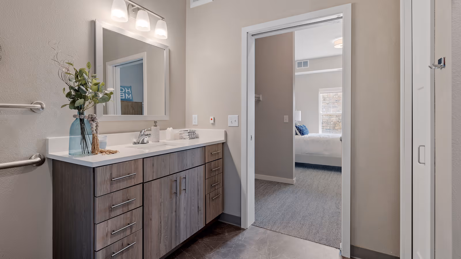 Modern bathroom vanity with sink, mirror, and decorative vase, opening into a bedroom through a doorway.