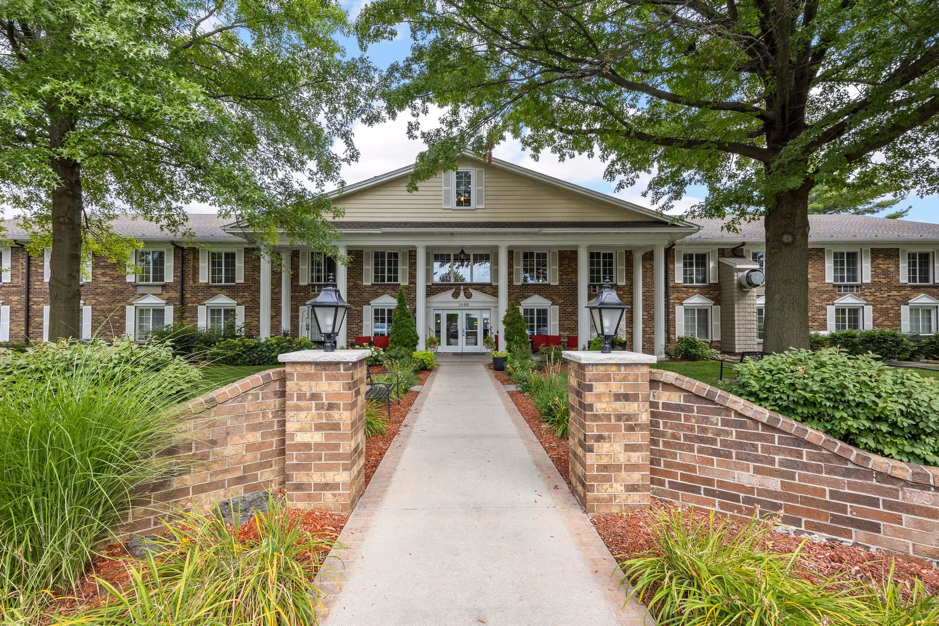 Front exterior view of a two-story senior living facility named Brown Deer Place, featuring a brick facade, white columns, a central entrance with glass doors, and a walkway flanked by brick walls and greenery under large trees.
