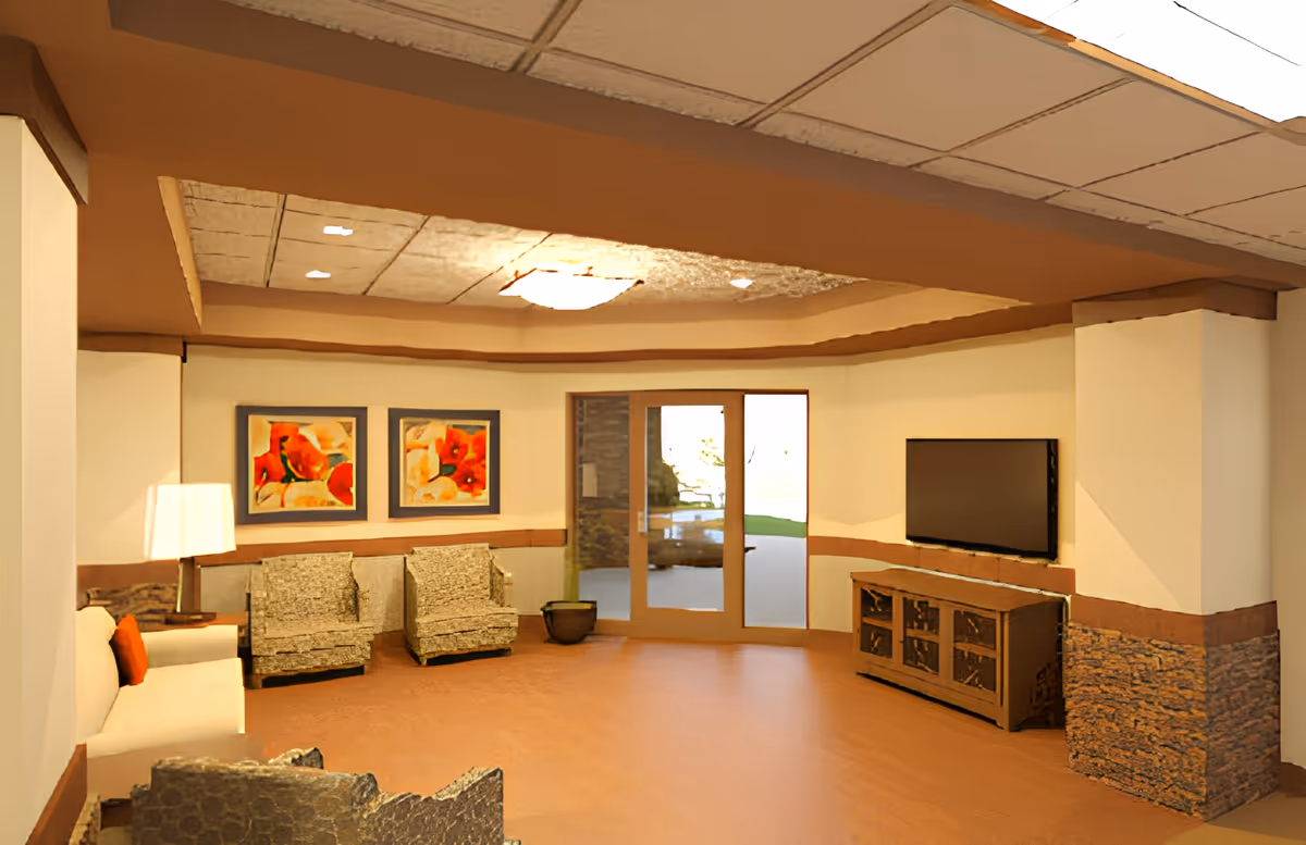 Bright communal lounge with chairs, a sofa, wall-mounted TV, artwork, and glass doors leading outside.