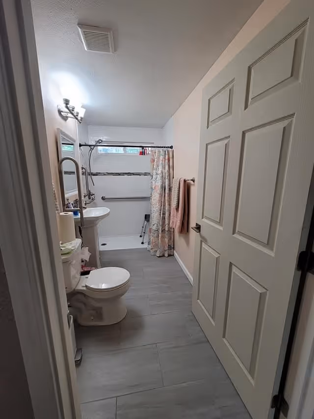 A clean bathroom with a white toilet, pedestal sink, and a walk-in shower with a floral shower curtain. There is a towel rack with a pink towel on the right wall and a mirror above the sink. The floor is covered with gray tiles and the walls are painted light beige.