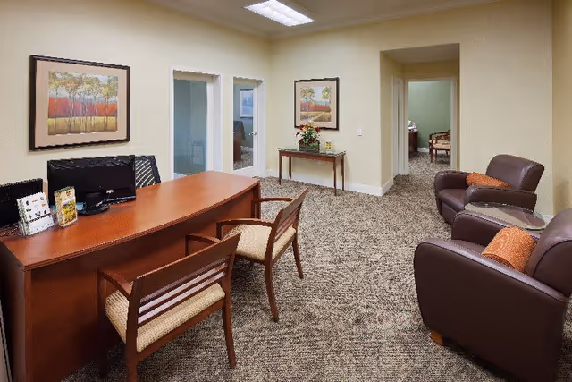 An office or reception area with a wooden desk and two chairs on one side. Behind the desk is a computer monitor. The room has beige walls with two framed paintings of trees. To the right, there are two brown leather armchairs with orange cushions and a small glass-top side table between them. A small wooden table with a flower arrangement is against the far wall, and there are two white doors with frosted glass panels. The carpet is patterned in neutral tones.