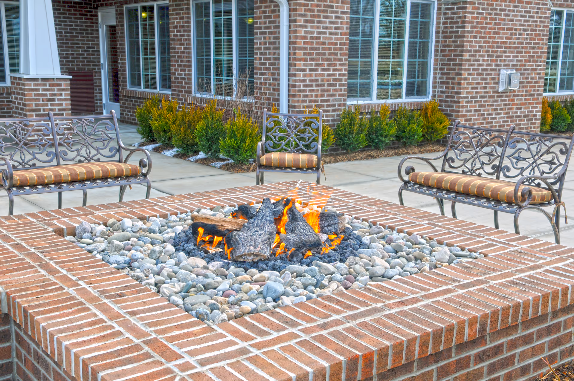 Outdoor seating area with a brick fire pit surrounded by decorative rocks and three metal benches with striped cushions, set in front of a brick building with windows and small shrubs.