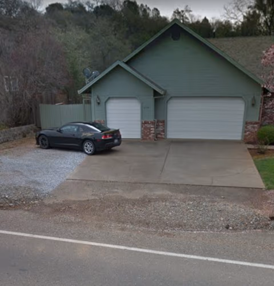 A green building with two white garage doors, a wide driveway, and a black car parked in front.