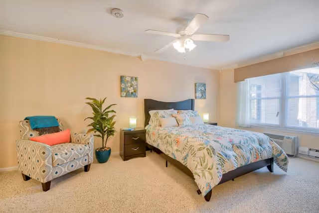 A bright bedroom with a large window covered by blinds and a beige valance. The room features a bed with a floral patterned comforter and multiple pillows, two dark wood nightstands with lamps, a patterned armchair with a coral pillow and teal throw, and a potted plant. The ceiling has a white fan with lights.