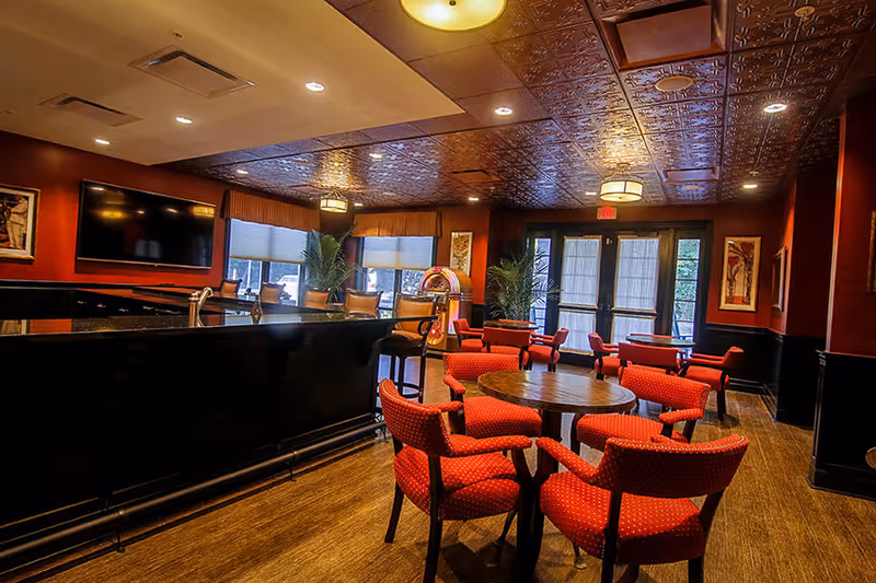 A warm, well-lit lounge with red upholstered chairs around round tables, a bar area, and a wall-mounted TV.
