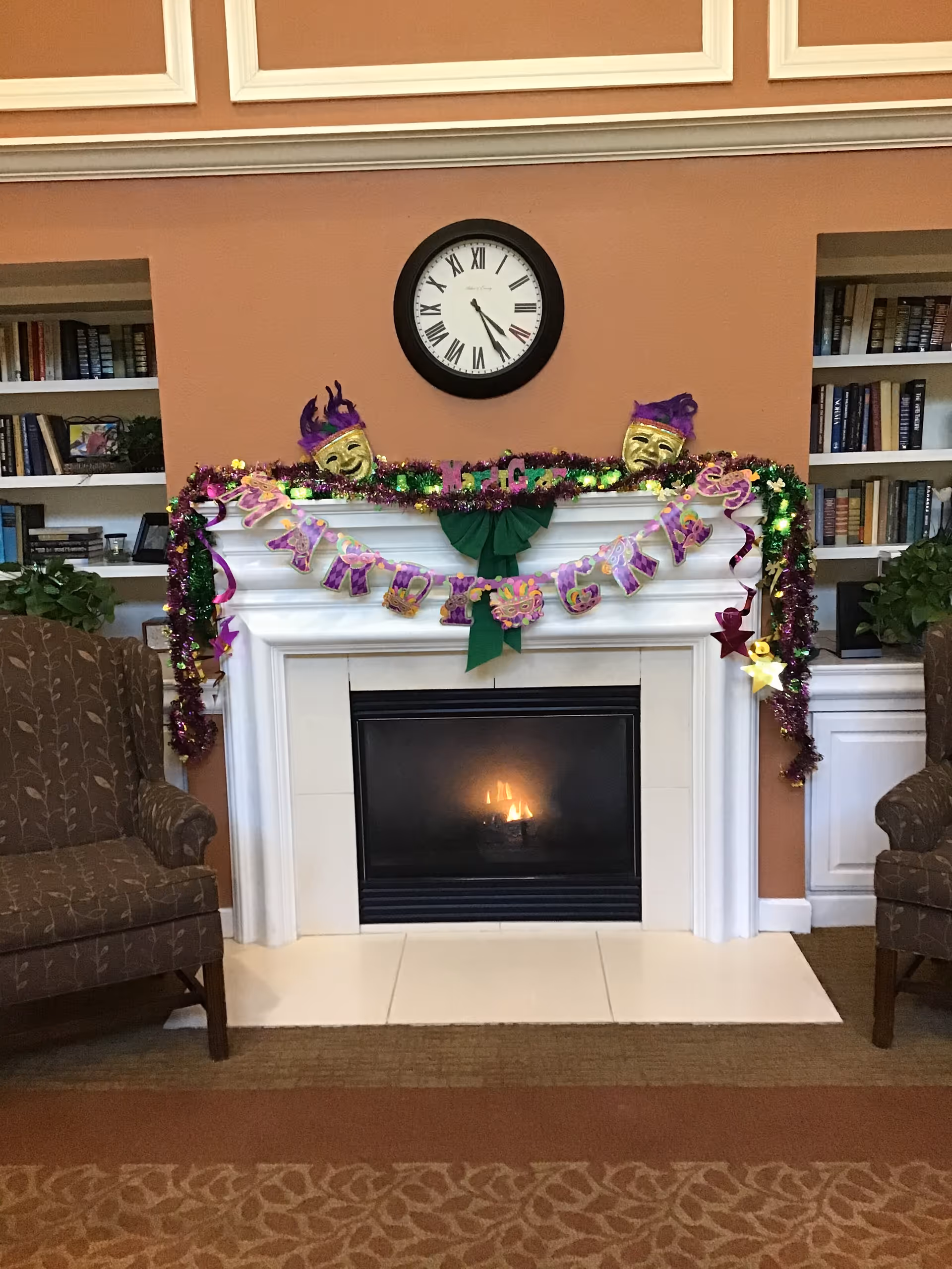 A cozy living room area featuring a white fireplace with a small fire burning inside. Above the fireplace is a round clock showing the time as 5:45. The mantel is decorated with Mardi Gras-themed garlands, including purple, green, and gold tinsel, two gold theatrical masks with purple accents, and a banner that reads 'Mardi Gras'. On either side of the fireplace are built-in bookshelves filled with books and some decorative items. Two brown patterned armchairs are positioned on each side of the fireplace, and there are green plants on the shelves.