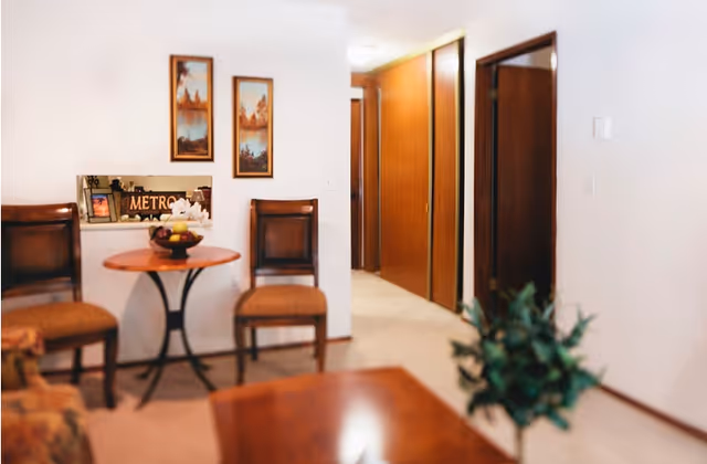 Interior view of a senior living facility showing a small round wooden table with a bowl of fruit on it, flanked by two wooden chairs with cushioned seats. The background features a hallway with wooden doors and two framed landscape paintings on the wall.