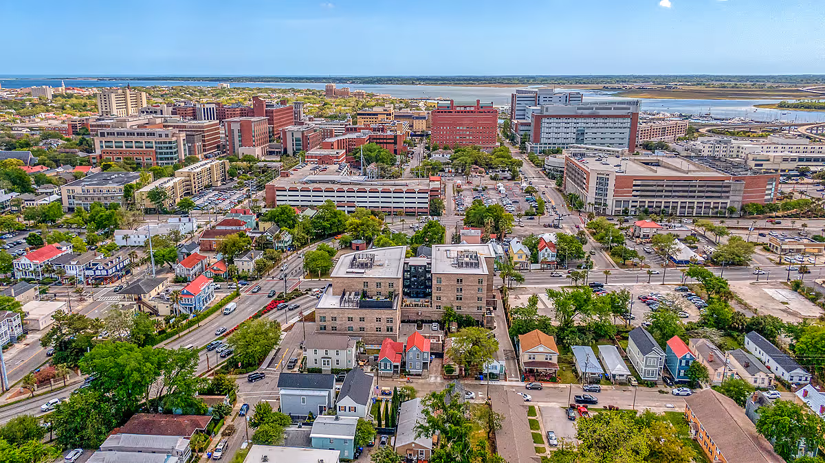 Aerial view of an urban area in Charleston showing a mix of residential houses, mid-rise buildings, parking lots, and greenery with a body of water visible in the background under a partly cloudy sky.