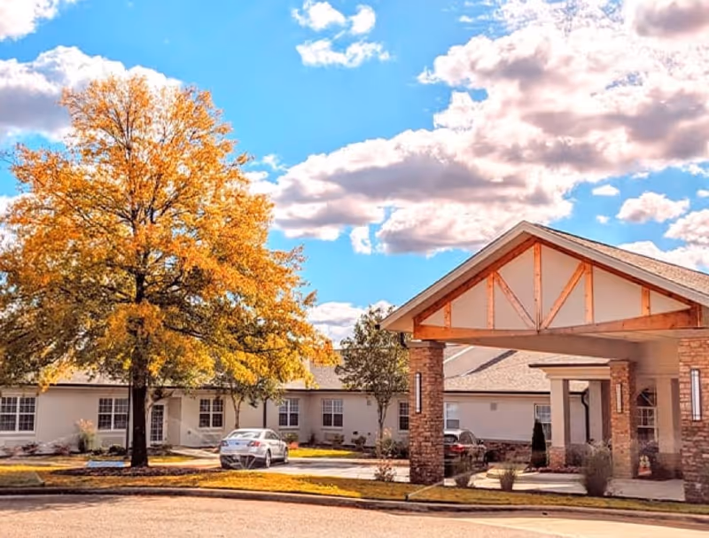 Front entrance of a single-story senior living building with a covered porte-cochere, parked cars, and a large tree with autumn foliage under a blue sky.