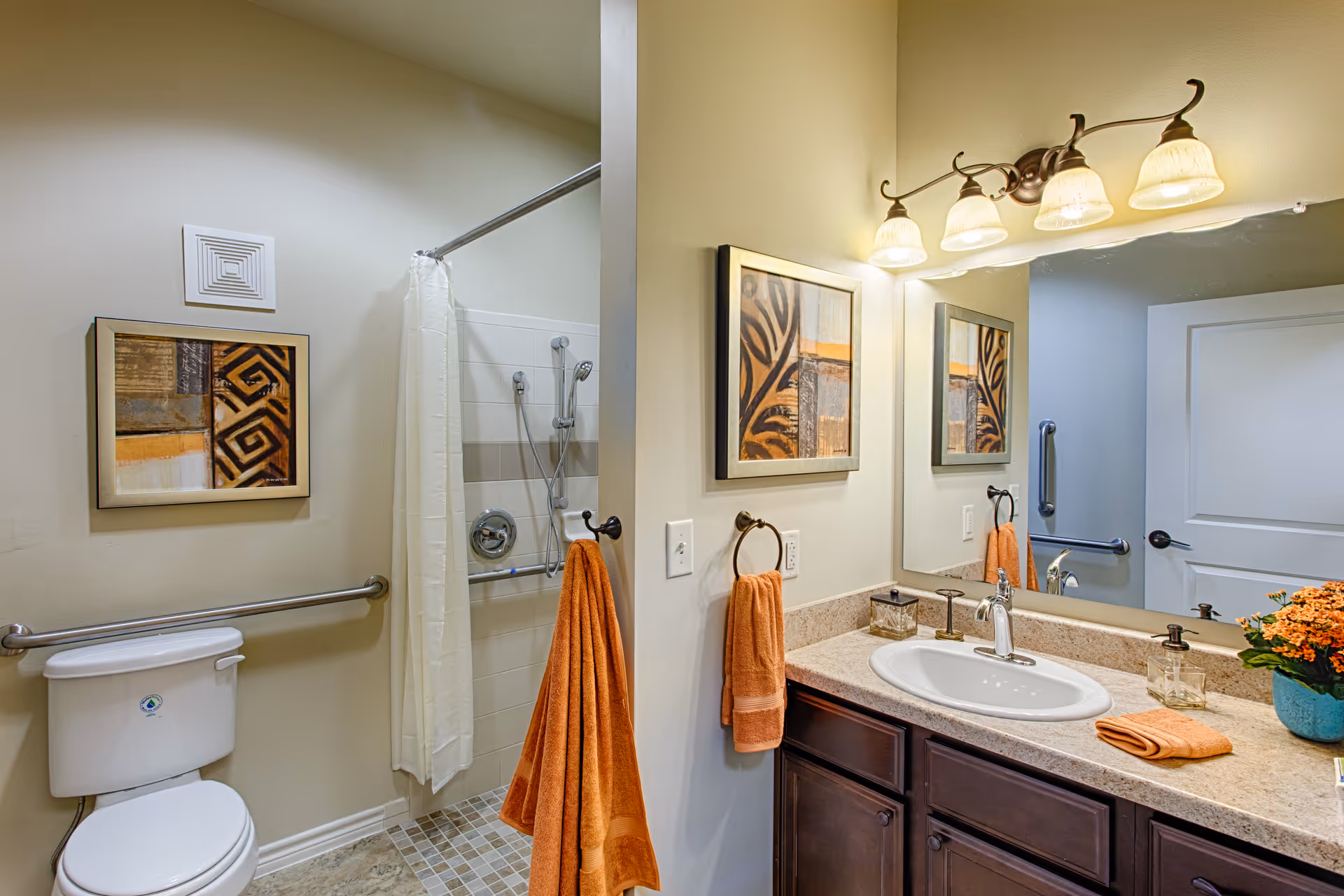 A clean and well-lit bathroom featuring a toilet with a safety grab bar, a walk-in shower with a curtain and handheld showerhead, a vanity with a sink, countertop, and dark wood cabinets. The bathroom has beige walls, a framed abstract artwork, orange towels hanging on towel rings, a large mirror above the sink, and a light fixture with four bulbs. A small potted plant with orange flowers is placed on the countertop.